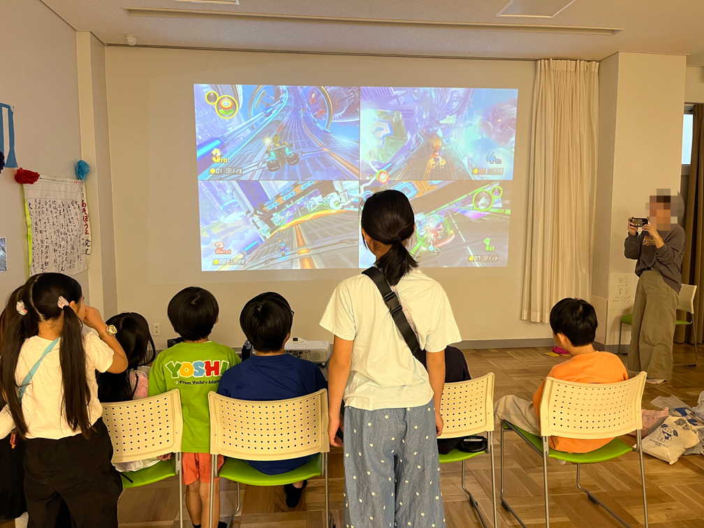 吉西コミセン祭りのカーレースゲーム大会で、壁に大きく映された4人分のレース画面を見る子どもたち タイトル